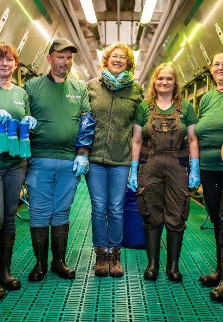 Das Bild zeigt f&uuml;nf Personen, vier Frauen und einen Mann, in einem modernen Kuh-Melkstand. Die Personen tragen Arbeitskleidung, darunter T-Shirts, eine Latzhose und Stiefel. Zwei Personen tragen blaue Schutzhandschuhe und halten Werkzeuge oder Produkte, d