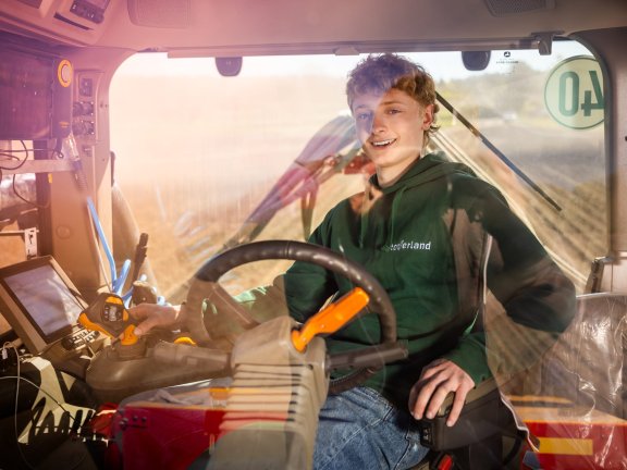 Ein Portr&auml;tfoto von einem jungen Mann mit lockigem braunem Haar, der einen gr&uuml;nen Kapuzenpullover und Blue Jeans tr&auml;gt, w&auml;hrend er in der Kabine eines modernen Traktors sitzt. Er l&auml;chelt freundlich in die Kamera und blickt &uuml;ber seine linke Schulter. Seine