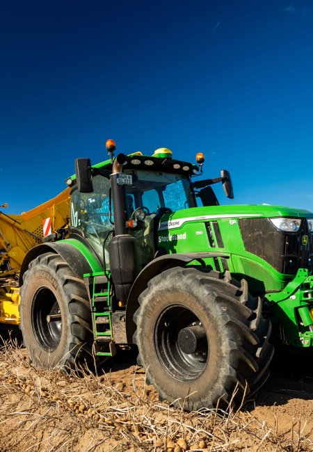 Ein gr&uuml;ner John-Deere-Traktor zieht einen gelben Kartoffelroder &uuml;ber einen abgeernteten Acker unter blauem Himmel.