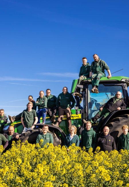 Ein gro&szlig;es Team von Landwirten und Mitarbeitern, etwa zwanzig Personen, posiert f&uuml;r ein Gruppenfoto in einem bl&uuml;henden gelben Rapsfeld an einem klaren, sonnigen Tag. Sie stehen und sitzen auf und um einen gro&szlig;en, gr&uuml;nen John Deere Traktor. Der Himmel ist