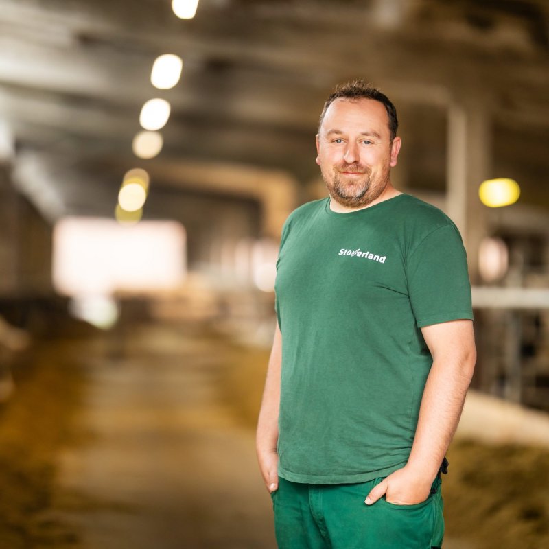 Ein Tierwirt in grüner Arbeitskleidung steht lächelnd im Kuhstall. Links im Bild fressen mehrere schwarz-weiße Kühe, der Hintergrund ist unscharf und zeigt die lange Stallgasse.