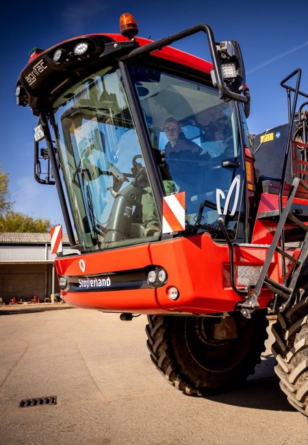Eine gro&szlig;e rot-schwarze, selbstfahrende Agrifac-Feldspritze steht auf einem gepflasterten Betriebshof vor Betriebsgeb&auml;uden.