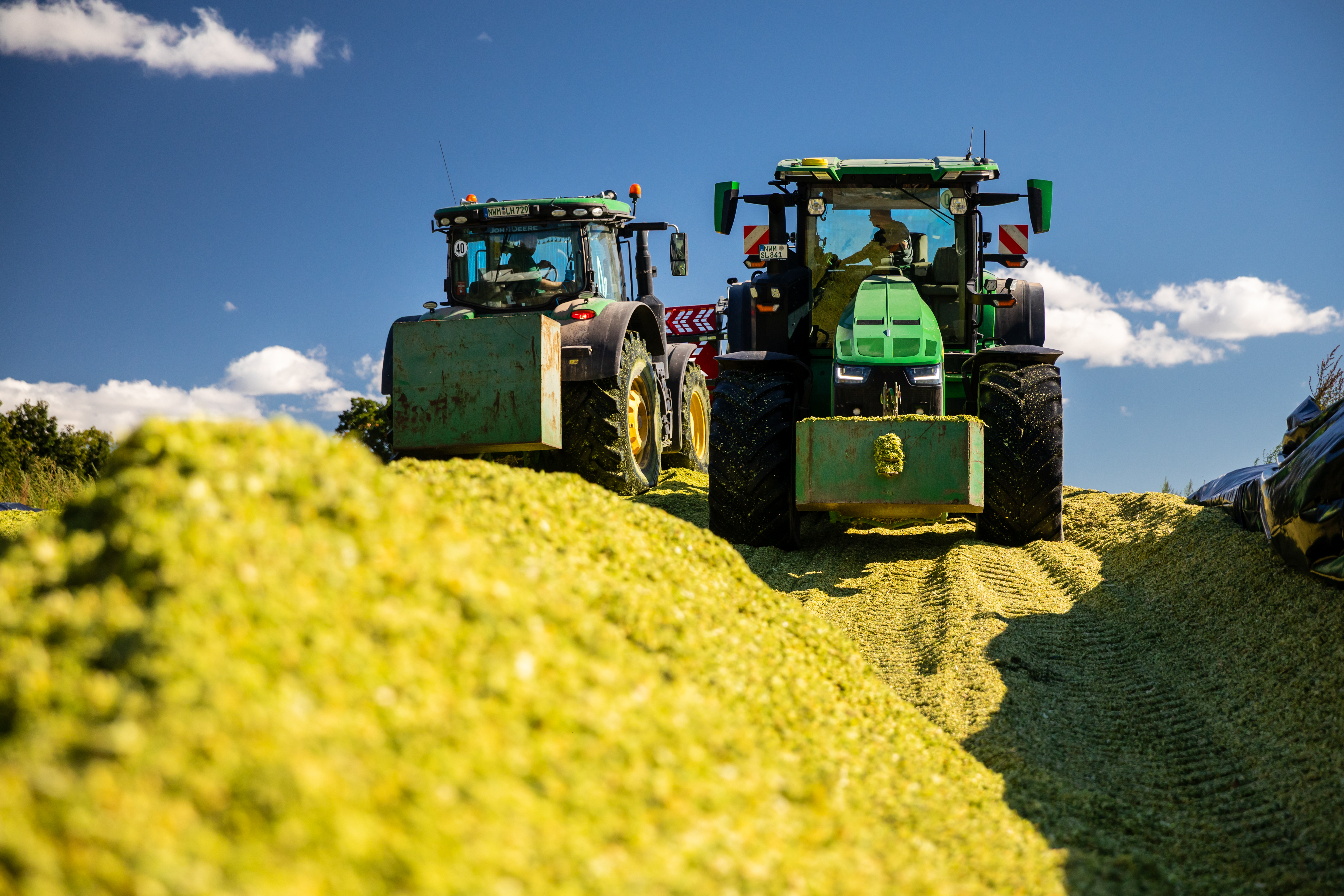 Zwei grüne Traktoren fahren hinter einem grünen Haufen Silage.