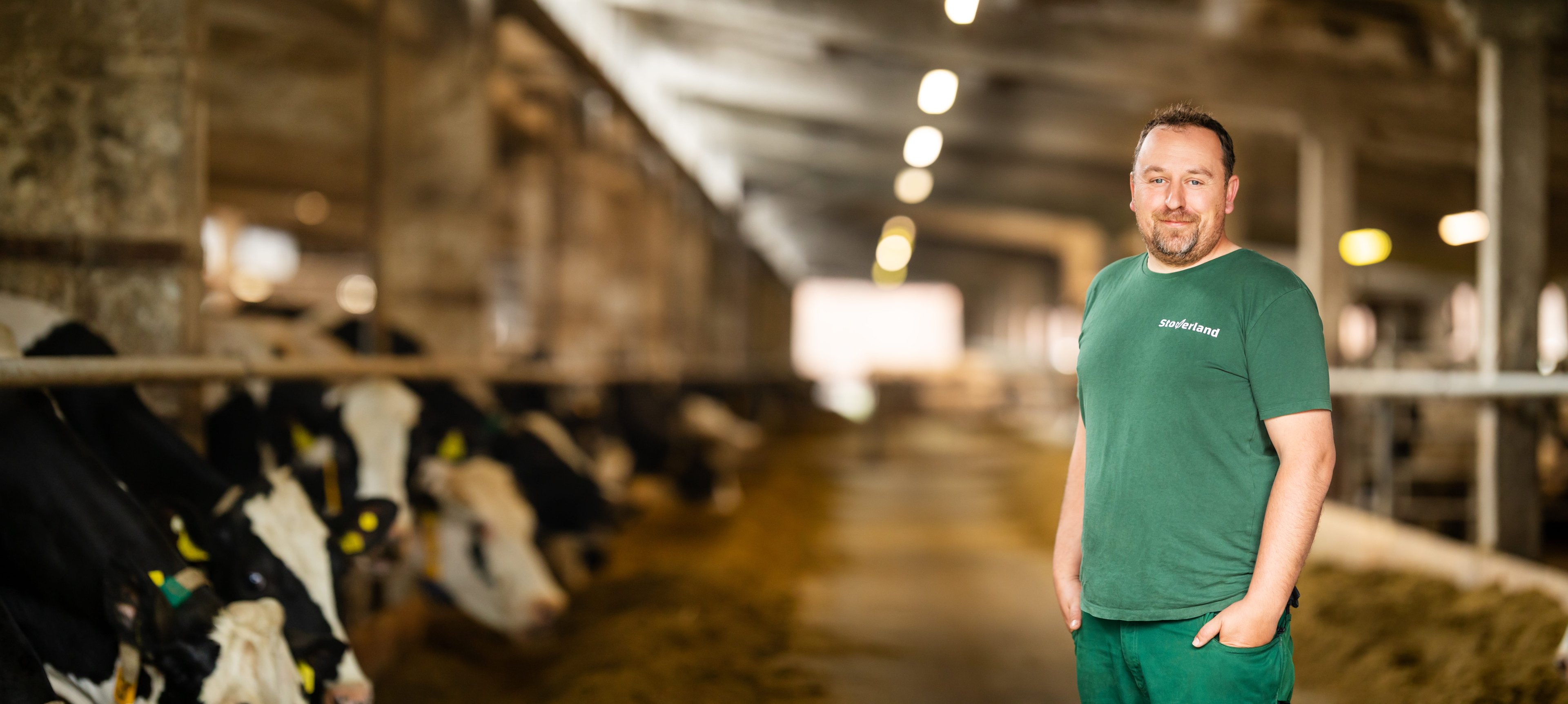 Ein Tierwirt in grüner Arbeitskleidung steht lächelnd im Kuhstall. Links im Bild fressen mehrere schwarz-weiße Kühe, der Hintergrund ist unscharf und zeigt die lange Stallgasse.